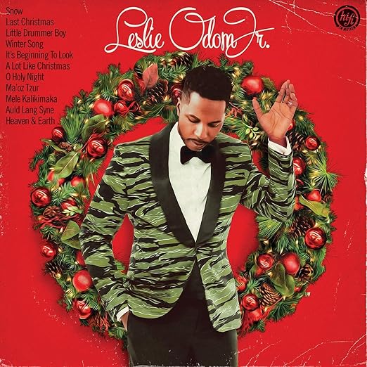 Image of the cover for "Leslie Odom Jr - The Christmas Album" by Tandem Coffee Roasters, featuring a well-dressed man in a green and black patterned suit jacket against a red background. A festive wreath surrounds him, and the artist's name, Leslie Odom Jr., is prominently displayed at the top with song titles like "Snow Last Christmas" to the left.