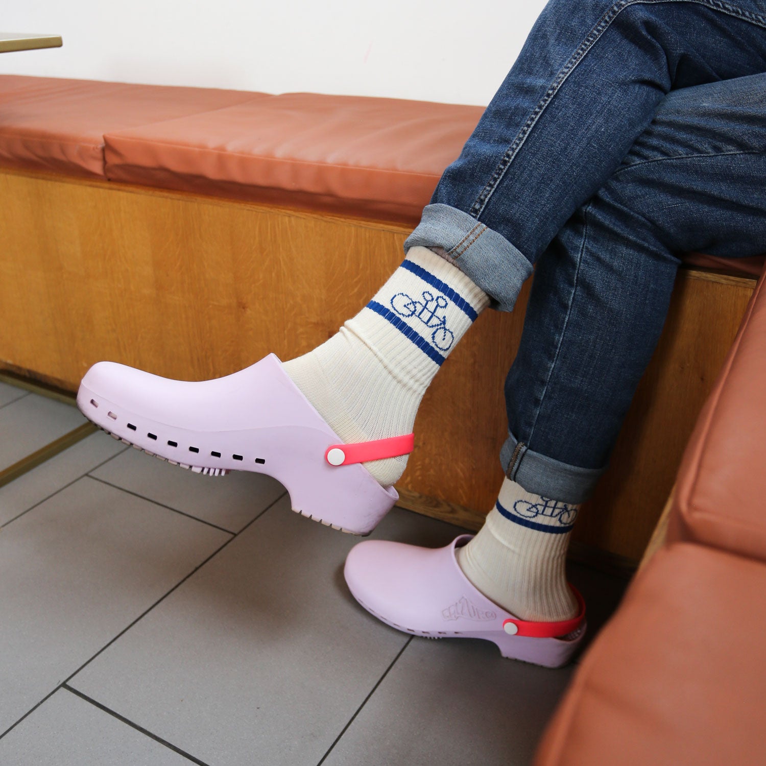 A person sits on a brown leather bench, wearing cuffed blue jeans and The Tandem Socks by Tandem Coffee Roasters, showcasing a blue bicycle design. Their outfit is complemented by lavender clogs with red straps. The aromatic scent of Tandem Coffee fills the air, enhancing the scene on the gray-tiled floor.