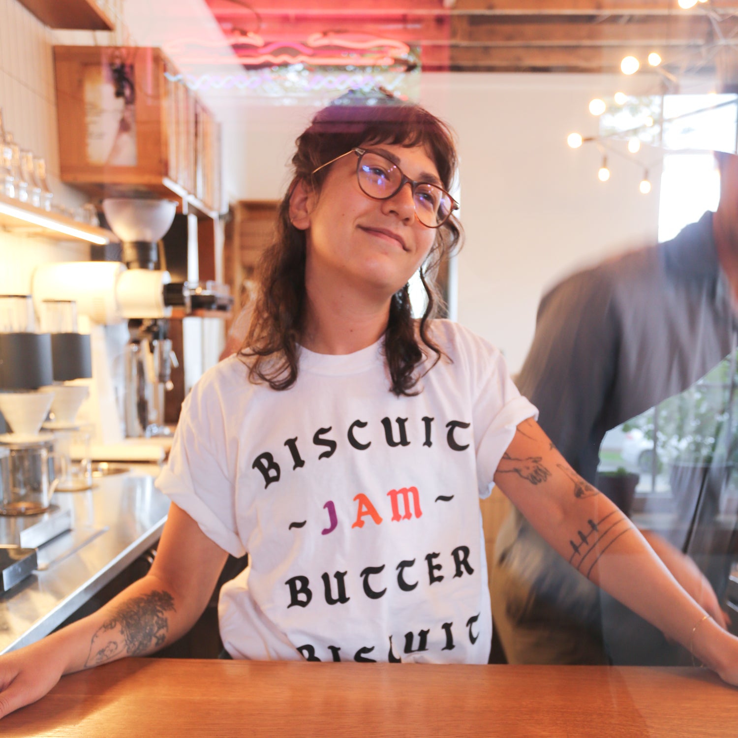 A smiling person with glasses, wearing a Tandem Coffee Roasters Loaded Biscuit tee reading "Best Biscuit Outside of the South," sitting at a cafe counter with a blurred server walking behind.