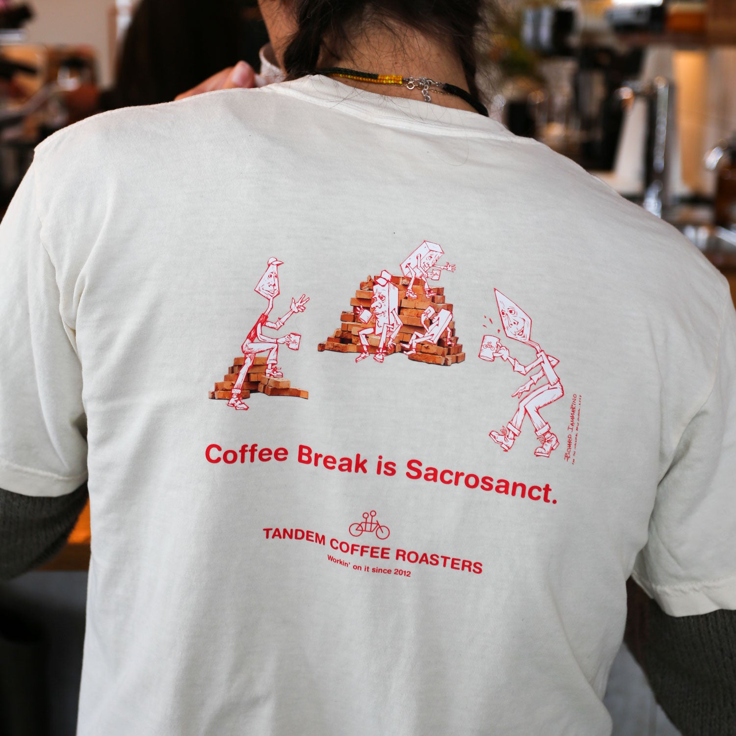 A person wearing the Brick Tee from Tandem Coffee Roasters, featuring red illustrations of characters stacking bricks, reminiscent of an issue from Brick Journal, and sipping coffee with the phrases "Coffee Break is Sacrosanct" and "Tandem Coffee Roasters.