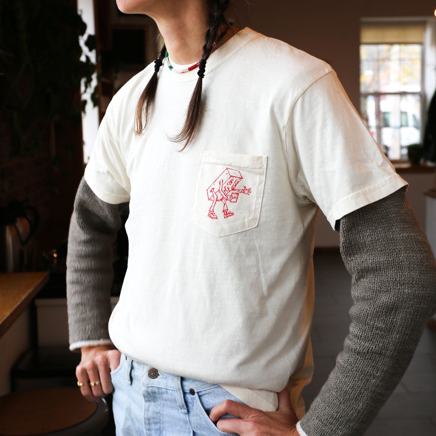 Wearing Tandem Coffee Roasters' Brick Tee, which features a striking red abstract design on the pocket, a person stands with hands on hips in a style reminiscent of Alexis Iammarino's scenes. They've layered the tee over a gray long-sleeved shirt. The warm glow of the cozy, plant-filled room behind them evokes pages from Brick Journal.