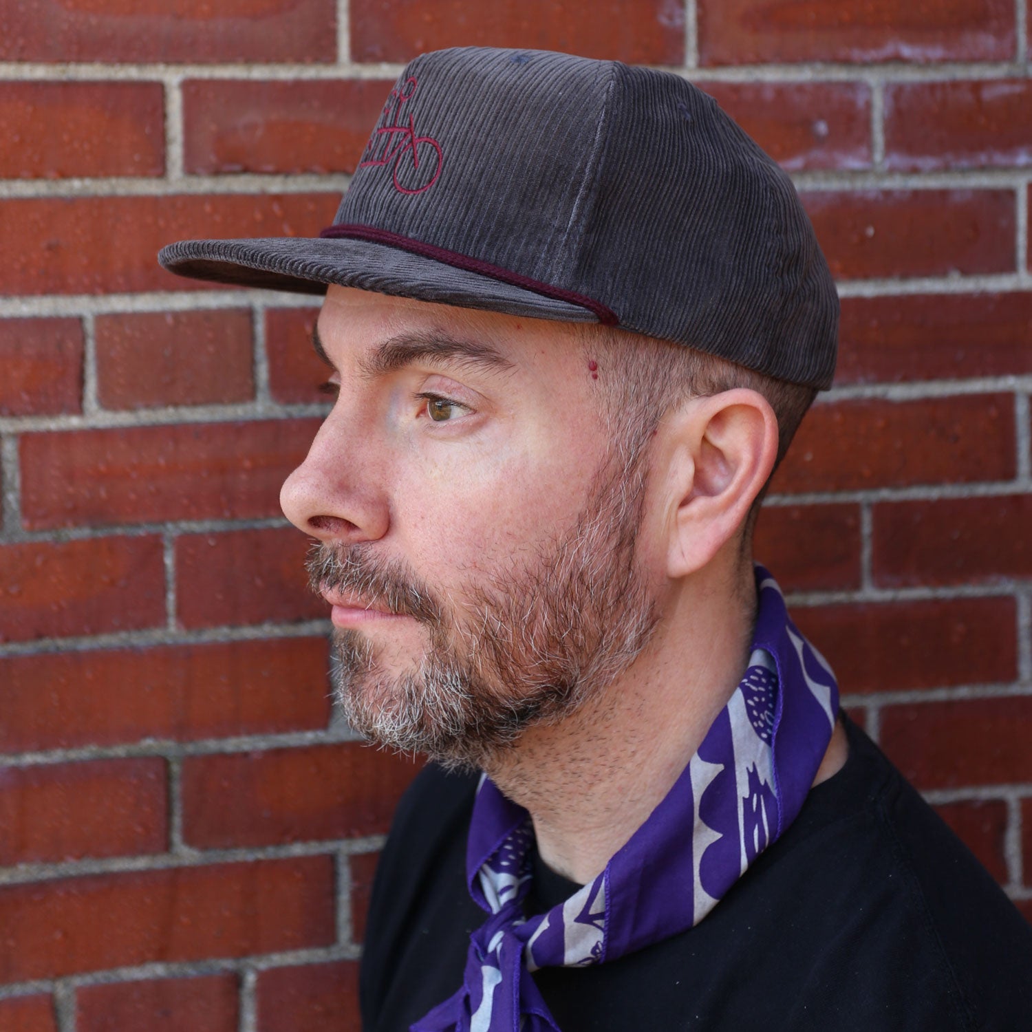 A bearded man in a dark cap and black shirt stands in profile against a brick wall, with a purple patterned scarf around his neck. His look is completed by a stylish Corduroy Tandem Hat from Tandem Coffee Roasters, adding an urban touch to the ensemble.