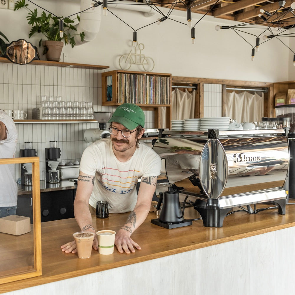 A barista with a mustache and green cap is smiling behind a wooden counter in a coffee shop, placing two drinks on the counter. The Tandem Coffee shop in Portland, Maine has a modern, minimalist interior with white tiles, exposed wooden beams, and various decorative items on shelves.