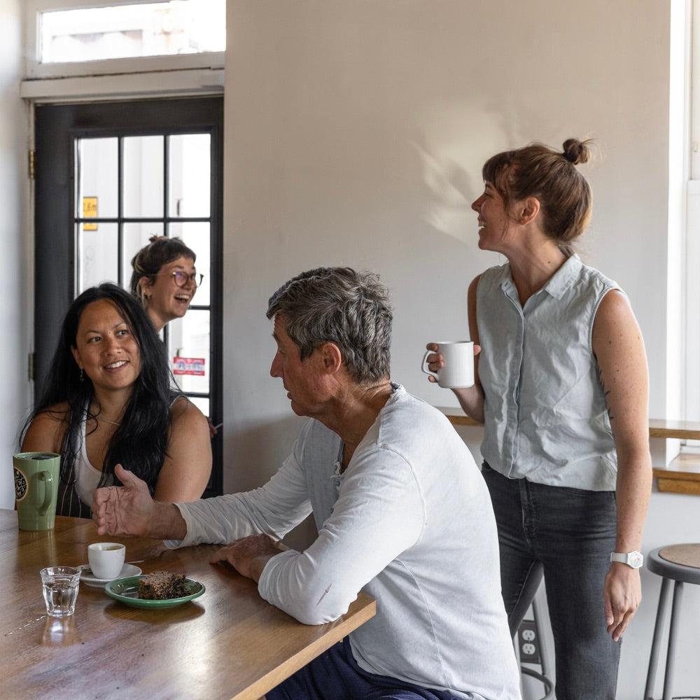 Four people are in a casual indoor setting, chatting and smiling. Two are seated at a table with coffee cups from Tandem Coffee, Portland Maine, while two others stand nearby. The room has light-colored walls and a windowed door in the background. The atmosphere is friendly and relaxed.