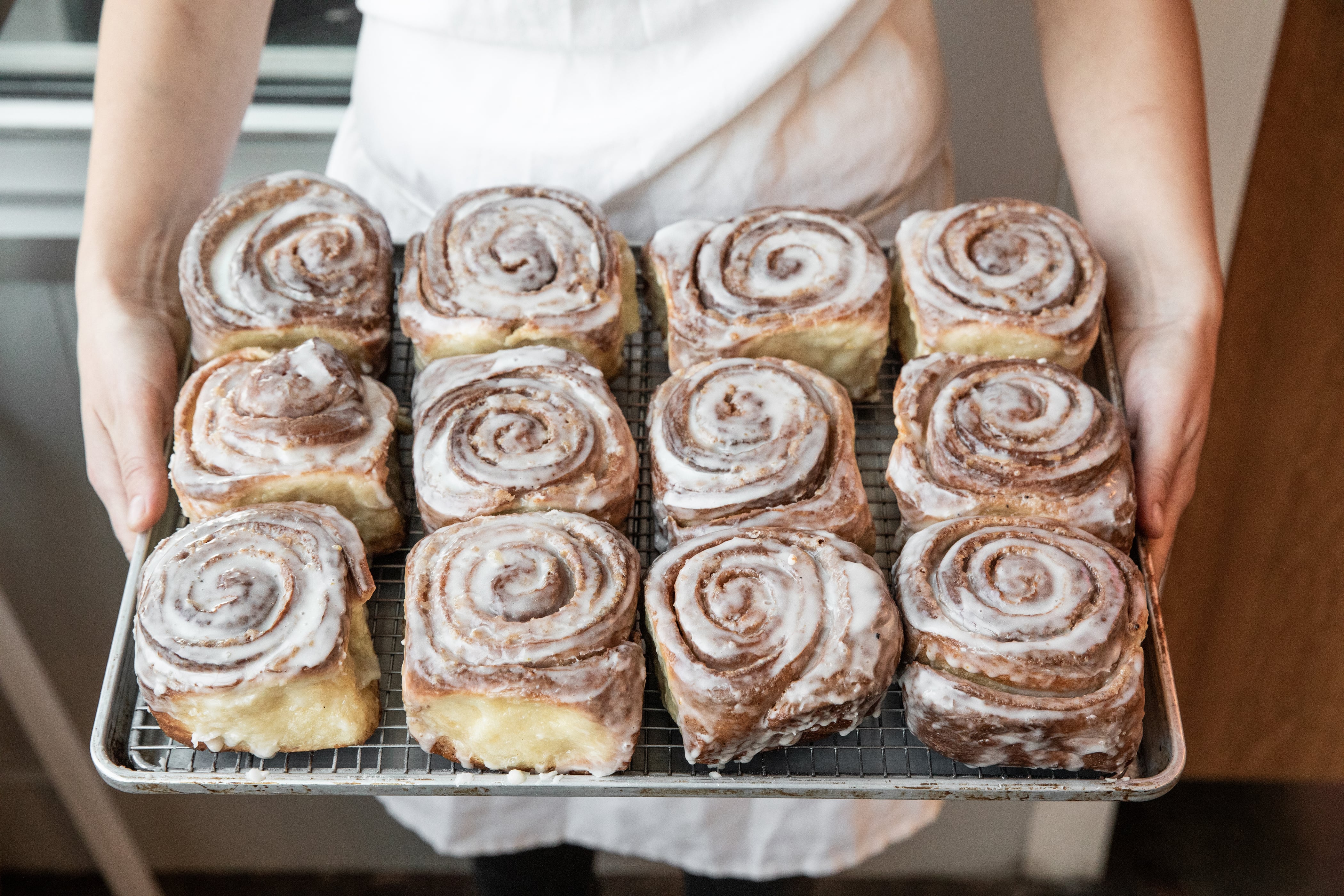 A person in a white apron holding a tray of freshly baked cinnamon rolls, each with a swirl pattern and glazed topping.