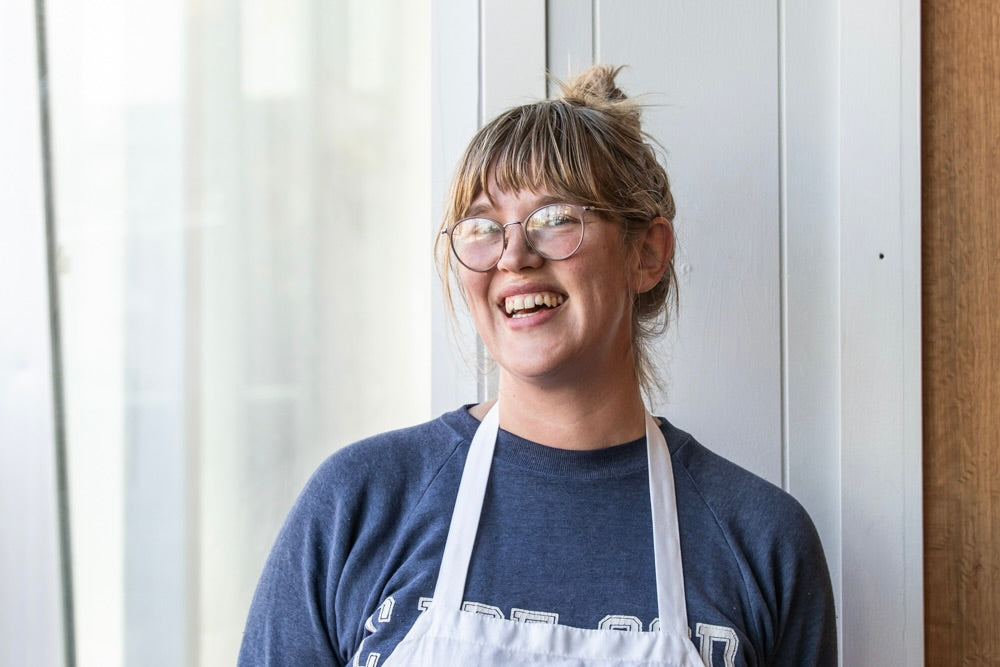 A person with blond hair tied up in a bun, wearing glasses and a navy blue sweatshirt under a white apron, is smiling brightly while standing in front of a white wall at Tandem Coffee in Portland, Maine.