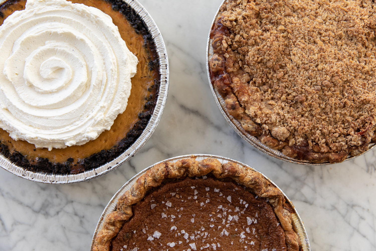 Top view of three pies: one with whipped cream swirl, one with crumb topping, and one sprinkled with coconut flakes, on a marble surface.