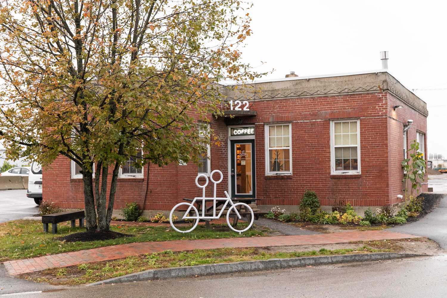 A small brick coffee shop in Portland, Maine, with "COFFEE" signage above the entrance and the number "122" displayed above the door. A tree with autumn leaves and a white metal sculpture resembling a tandem bicycle are in front of the shop. A bench and small garden are nearby.