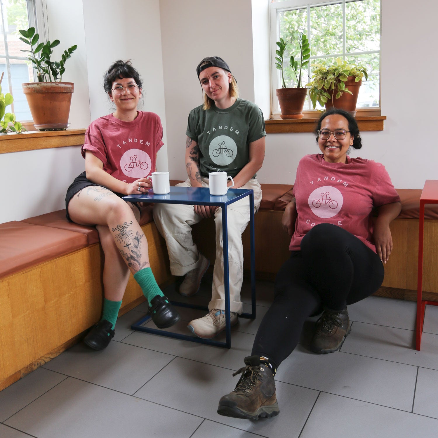 Three people sitting at a small table in a cozy café, smiling at the camera. They are wearing 100% cotton Moss Circle Logo Tees from Tandem Coffee Roasters in different colors, including MOSS color, and are surrounded by potted plants on a windowsill. Two of them have mugs in front of them, and two have visible tattoos.