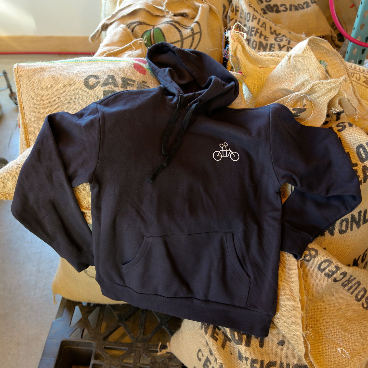 The Letter Pullover Hoodie in a super soft black fabric, featuring a bicycle design, is artfully displayed on burlap coffee sacks. The rustic setting, reminiscent of a cozy coffee shop or warehouse, is gently illuminated by natural light, enhancing the inviting textures and ambiance associated with Tandem Coffee Roasters.