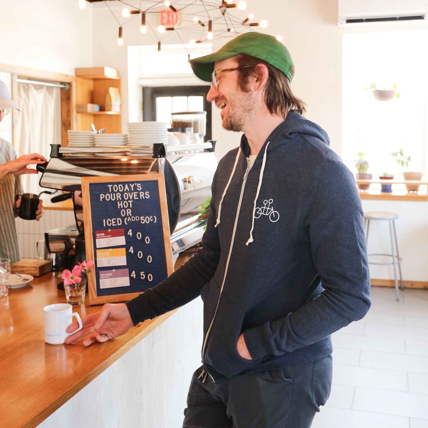 A man with glasses and a green cap sits on a brown bench against a plain white wall. He is wearing a cozy navy blue Letter Hoodie by Tandem Coffee Roasters with white drawstrings and a logo of a bicycle on the chest, along with gray shorts. His hands are clasped in his lap, and he is looking at the camera.