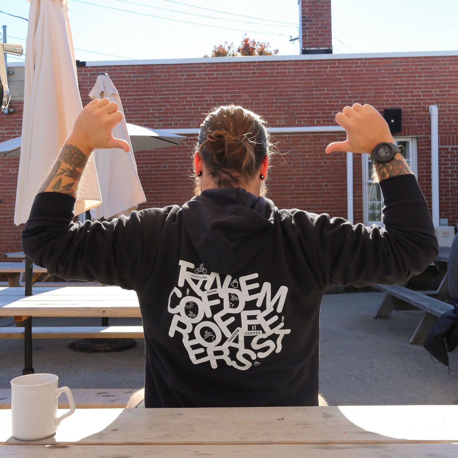 A person with tattoos and a bun is wearing a Letter Pullover Hoodie from Tandem Coffee Roasters, featuring bold text, while sitting at an outdoor table pointing at the design on the back. They have a watch on their wrist, and a coffee mug sits in front of them. In the background are brick walls and umbrellas.