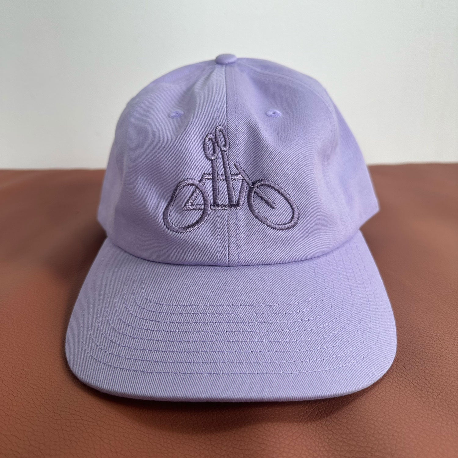 A Lilac Logo Dad Hat from Tandem Coffee Roasters rests on a brown surface. The adjustable hat features an embossed outline of a bicycle on the front panel. The plain white background highlights the cap's color and design.