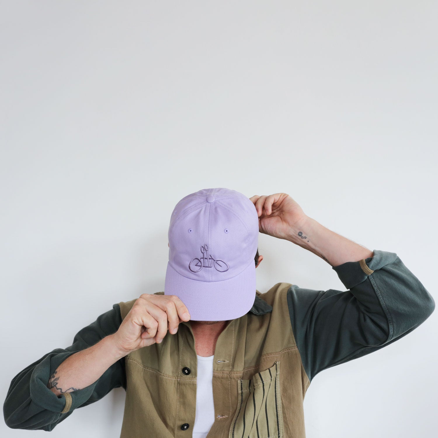 A person wearing a two-toned green jacket and a white shirt adjusts a Lilac Logo Dad Hat from Tandem Coffee Roasters made of 100% cotton with an embroidered bicycle logo. The person’s face is mostly obscured by the hat, and they have a small tattoo on their right wrist. The background is plain white.