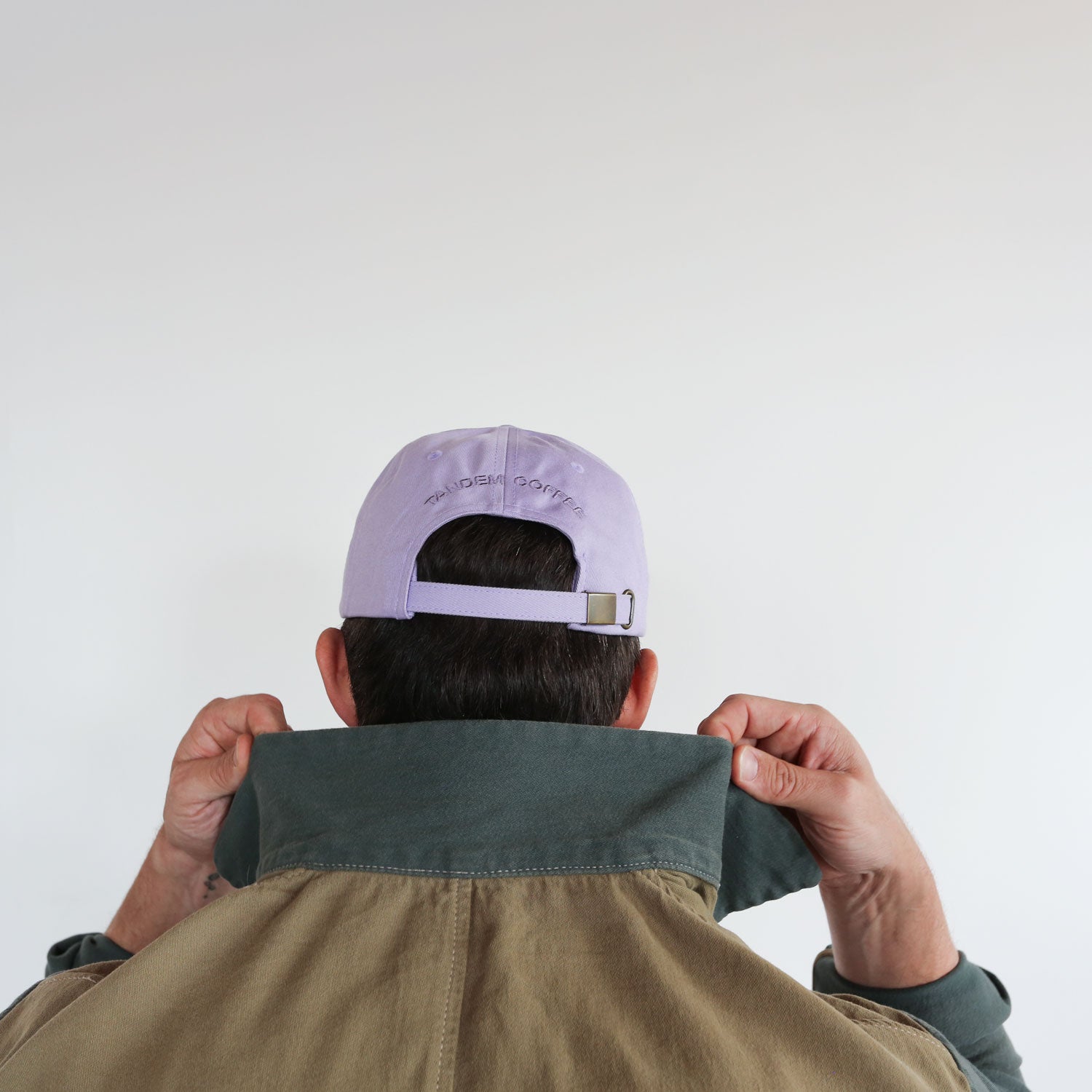 A person with short dark hair is seen from behind, raising the collar of their green jacket with both hands. They are wearing a light purple Lilac Logo Dad Hat by Tandem Coffee Roasters made from 100% cotton, featuring a small buckle on the back, against a plain white background.