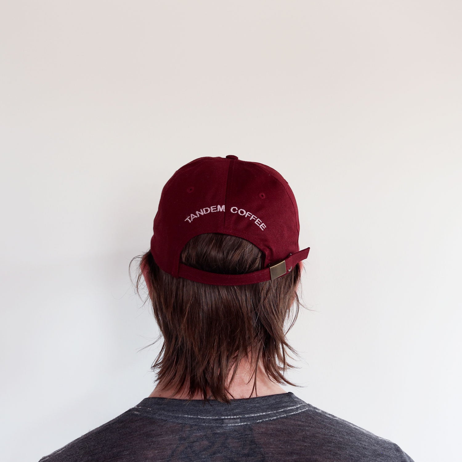 A person wearing a maroon, 100% cotton Maroon Logo Dad Hat from Tandem Coffee Roasters adjusts it, their face partially obscured. They have tattoos on both arms and are dressed in a grey t-shirt. The background is a plain white wall.