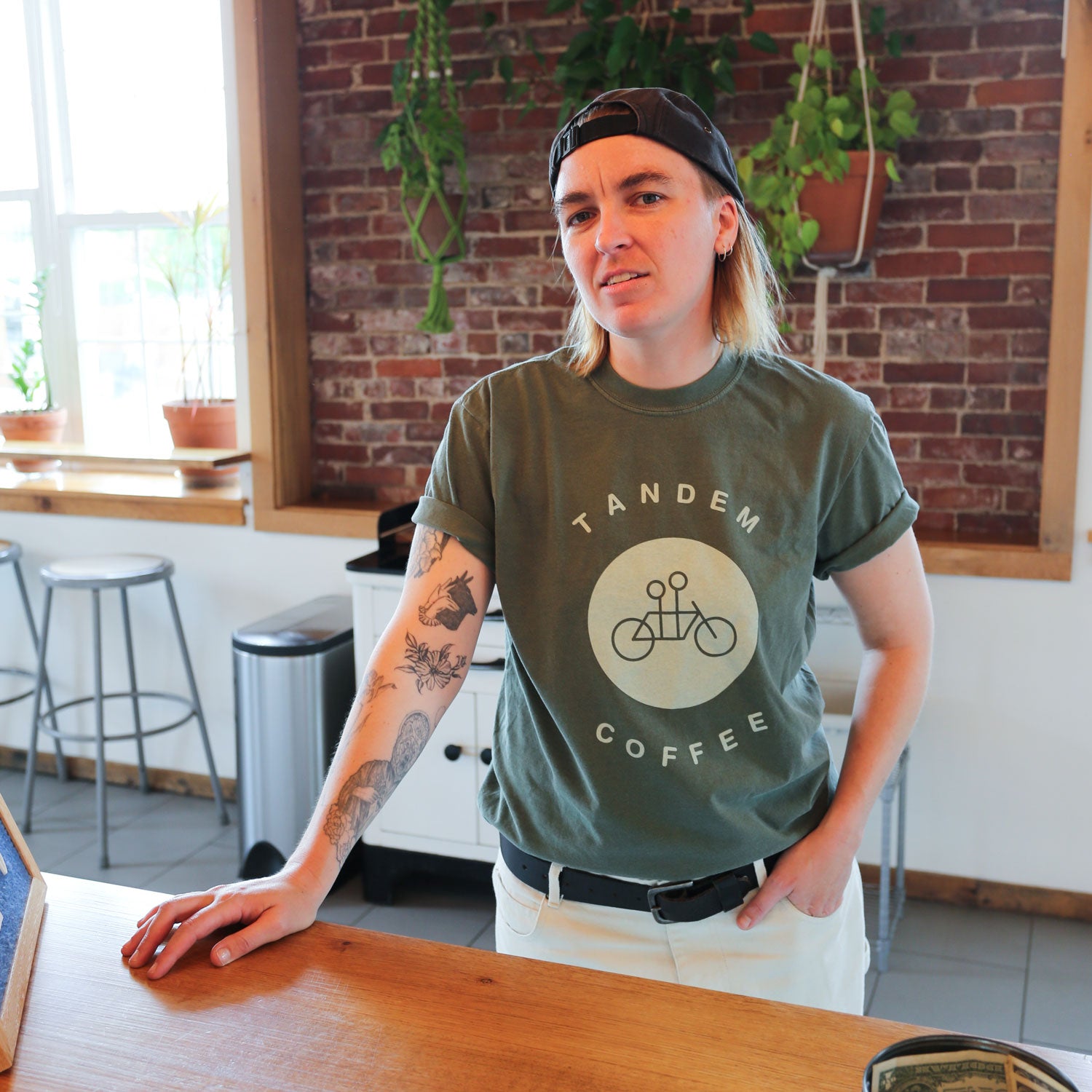 A person with shoulder-length hair and a backward baseball cap stands behind a wooden counter. They are wearing a green "Moss Circle Logo Tee" from Tandem Coffee Roasters made from 100% cotton for ultimate comfort. Tattoos adorn one arm, and the background features brick walls, hanging plants, and some stools.