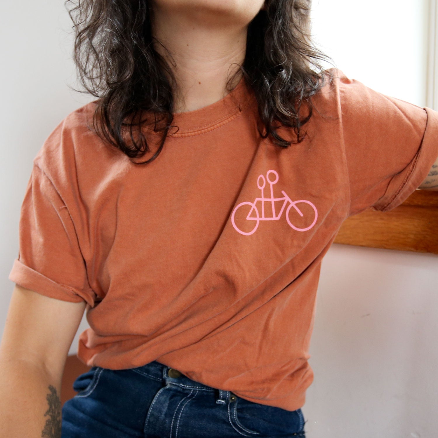 A person with long, wavy hair and wearing glasses stands against a plain background, looking slightly upwards and smiling. They are wearing a casual elegant, orange cotton Tandem Coffee Roasters Bike + Tandem Coffee Tee with a pink bicycle graphic and have tattoos on their left arm. Their hands are behind their back.