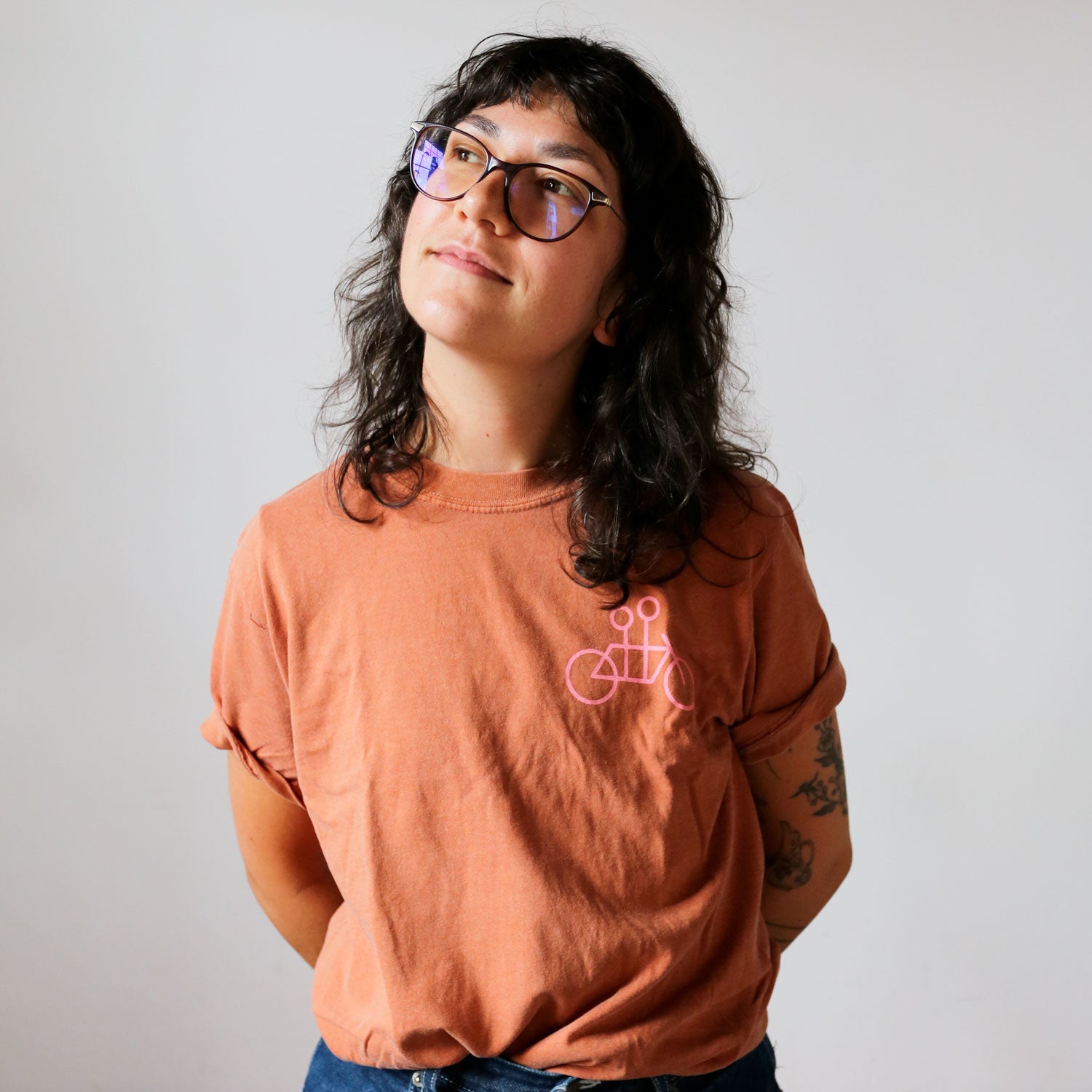 A person with long, wavy hair and wearing glasses stands against a plain background, looking slightly upwards and smiling. They are wearing a casual elegant, orange cotton Tandem Coffee Roasters Bike + Tandem Coffee Tee with a pink bicycle graphic and have tattoos on their left arm. Their hands are behind their back.