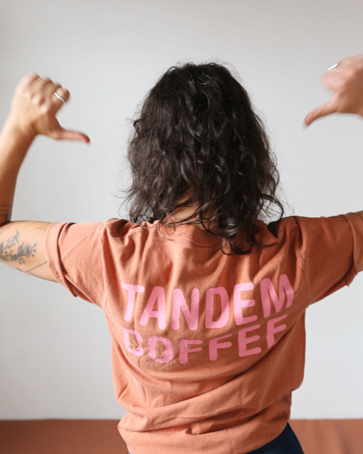 A person with curly hair, seen from behind, is wearing a casual elegant brown Bike + Tandem Coffee Tee from Tandem Coffee Roasters with the words "Tandem Coffee" printed in pink. They have their thumbs pointed towards themselves. The person has tattoos on their arm. The background is plain and out of focus, highlighting the logo tee's cotton Comfort Color fabric.