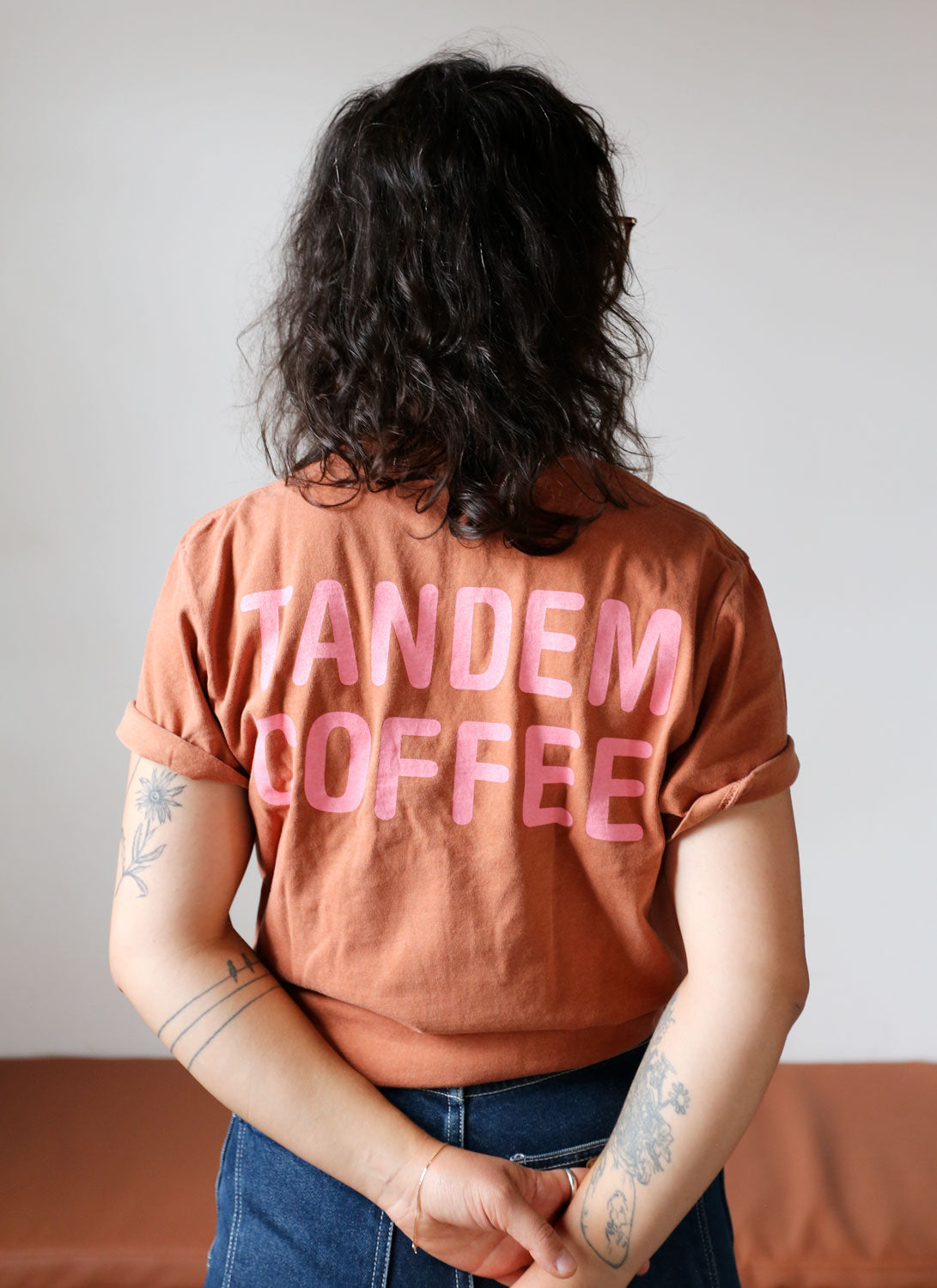 A person with shoulder-length wavy hair, wearing one of the Bike + Tandem Coffee Tees from Tandem Coffee Roasters in brown with the words "Tandem Coffee" in pink letters, stands with their back facing the camera. Their arms are crossed behind their back, revealing several tattoos on their left arm.