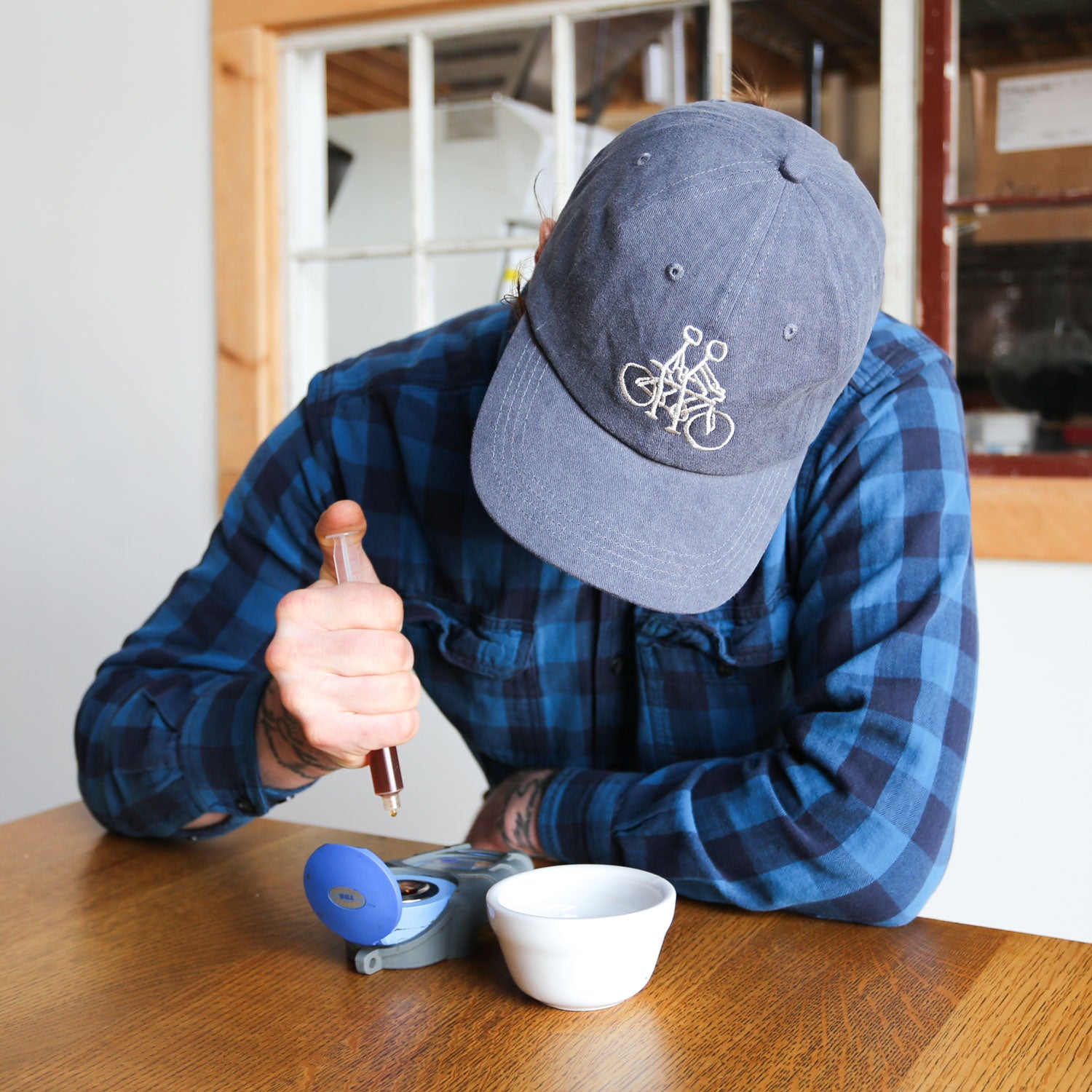 Wearing a Russ Pope Tandem Hat from Tandem Coffee Roasters, a person in a blue flannel shirt leans over a table, using a syringe-like tool to extract liquid from a small blue device next to a white bowl in an indoor setting with creative flair. A window is seen in the background.