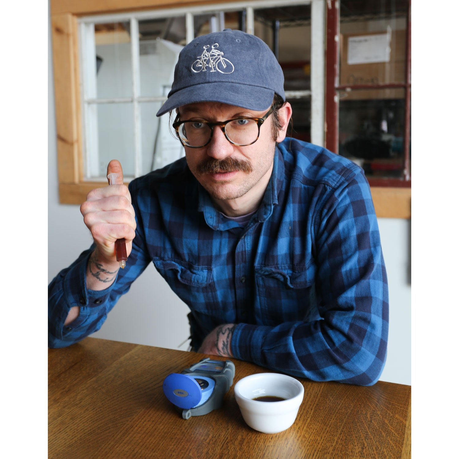 A mustached individual with glasses, sporting a blue plaid shirt and a Russ Pope Tandem Hat from Tandem Coffee Roasters, holds a dropper over a digital refractometer. Beside them on the wooden table is a cup of coffee, with multi-paned windows creating an artistic backdrop.