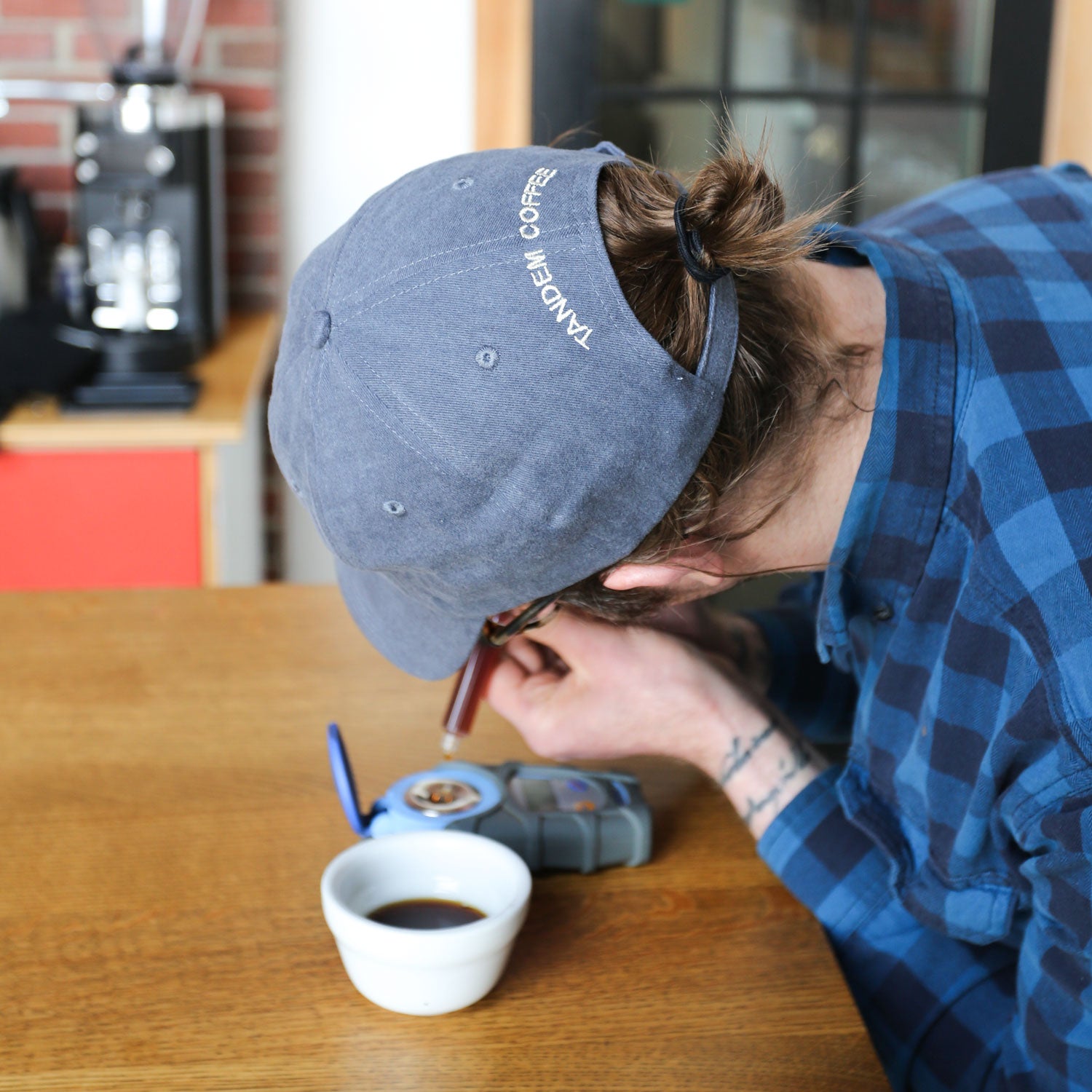 A person wearing a Russ Pope Tandem Hat and blue plaid shirt leans over a table, closely observing something with a magnifying glass. Nearby, there's a cup of coffee and a sleek coffee machine backdrop from Tandem Coffee Roasters.