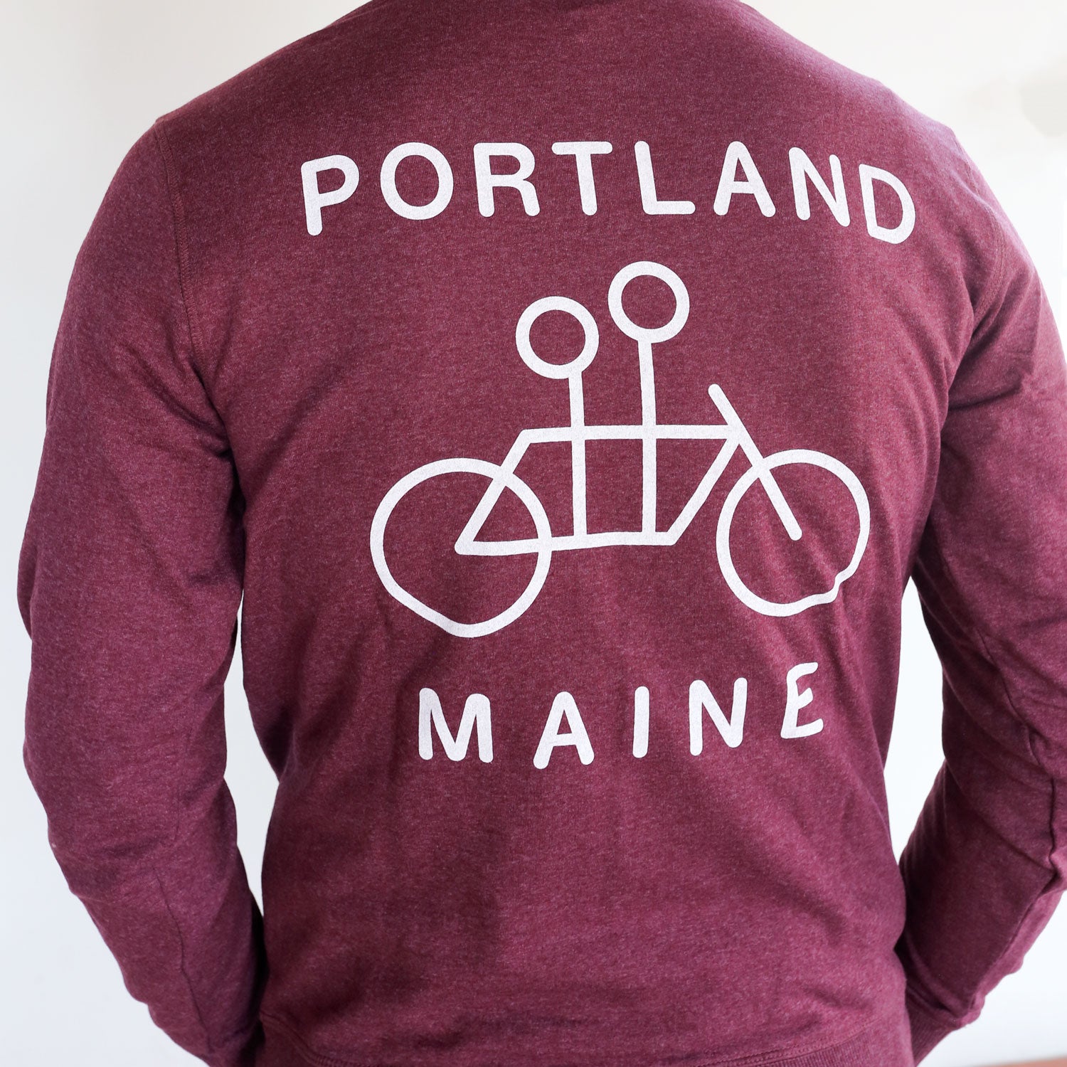 A person is wearing a Maroon Recycled Pullover Hoodie by Tandem Coffee Roasters with "portland maine" in white letters and a simple white bicycle graphic below the text, against a plain background.