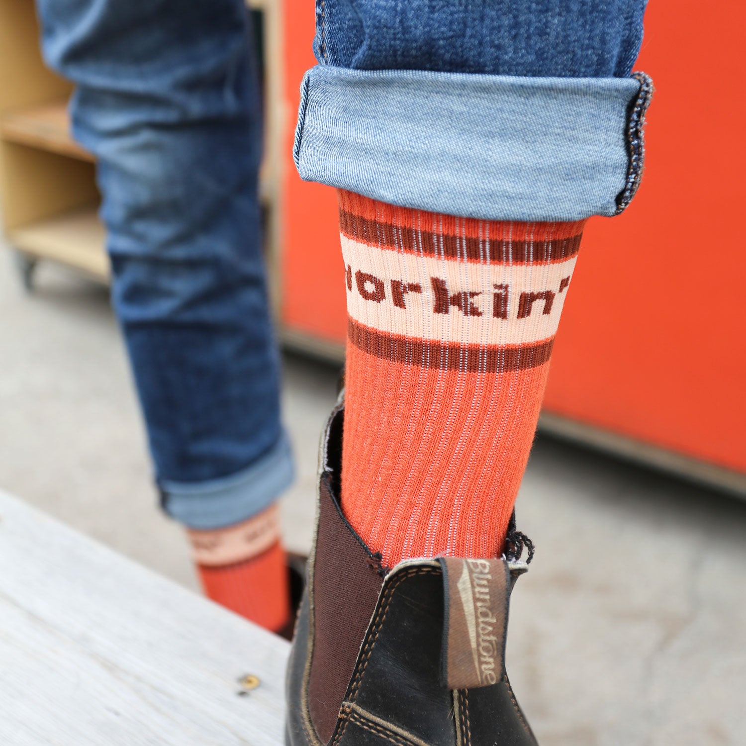 Close-up of a person's lower legs in rolled-up jeans, wearing cozy "The Tandem Socks" by Tandem Coffee Roasters and brown Blundstone boots. A red wall and wooden shelf set the backdrop, creating a scene that pairs perfectly with a freshly brewed cup from Tandem Coffee.