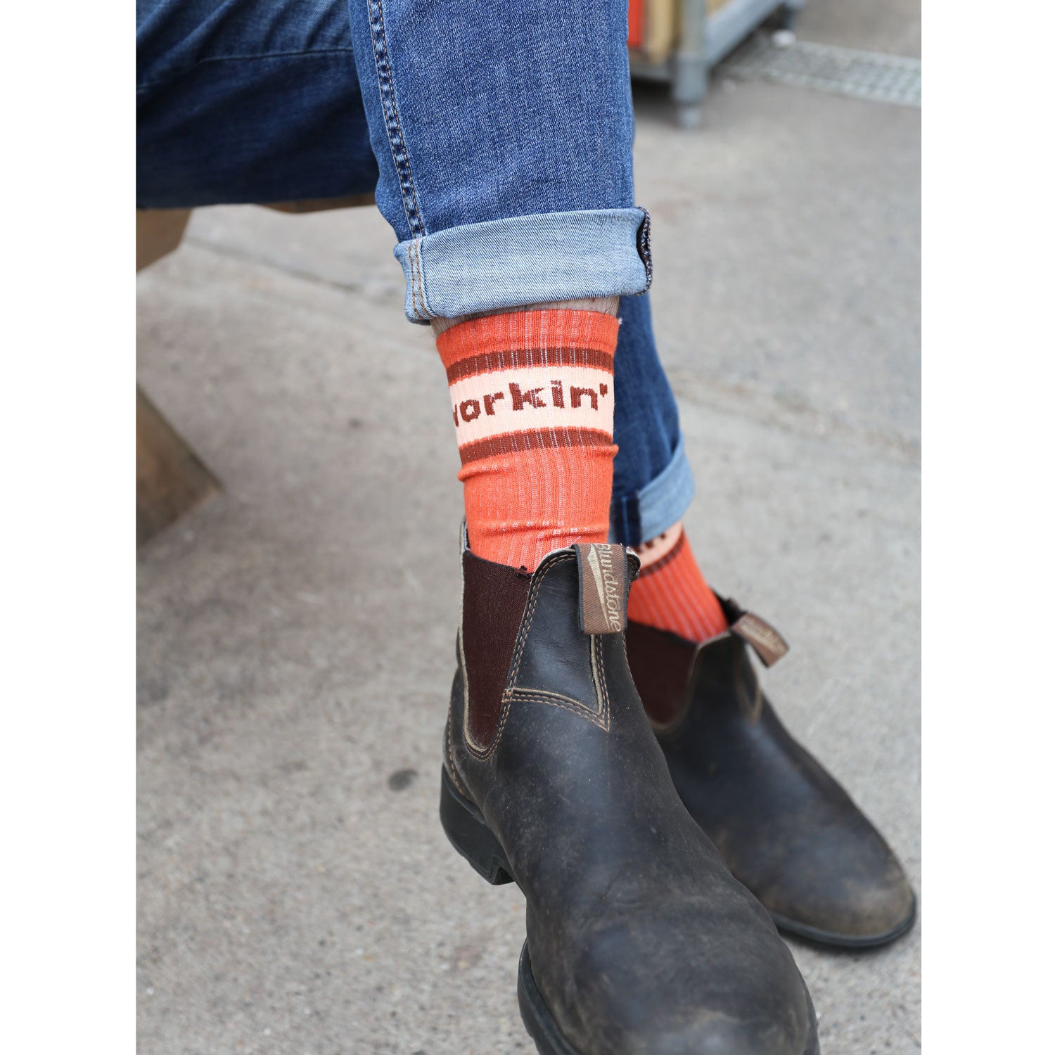 A person is seated wearing cuffed blue jeans and brown leather boots. Their cotton socks, named "The Tandem Socks" by Tandem Coffee Roasters, are vibrant in orange and feature the word "Workin’," adding a casual touch to the outfit. Nearby, a cup from Tandem Coffee rests on the textured pavement, completing the relaxed scene.