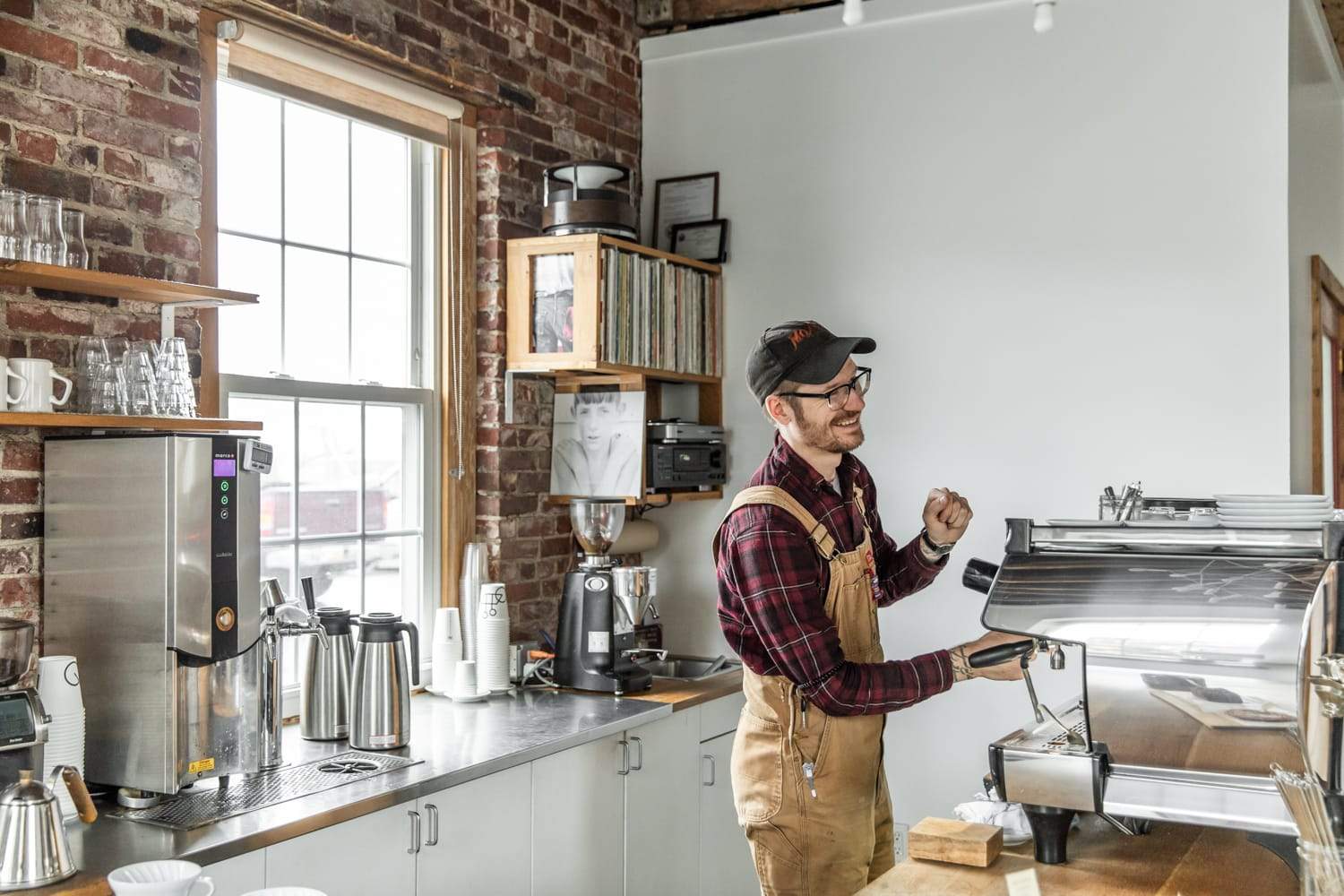 A cheerful barista in a cap and apron making coffee in a cozy, brick-walled cafe with modern equipment and sunny windows.