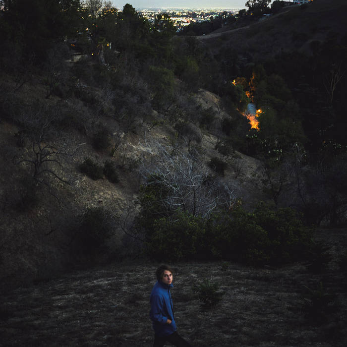 A man in a blue shirt sits pensively on a grassy hillside during twilight, with sparsely vegetated slopes behind him and distant city lights glowing beneath a dusky sky. He quietly listens to "Kevin Morby - Singing Saw" while sipping on Tandem Coffee Roasters.