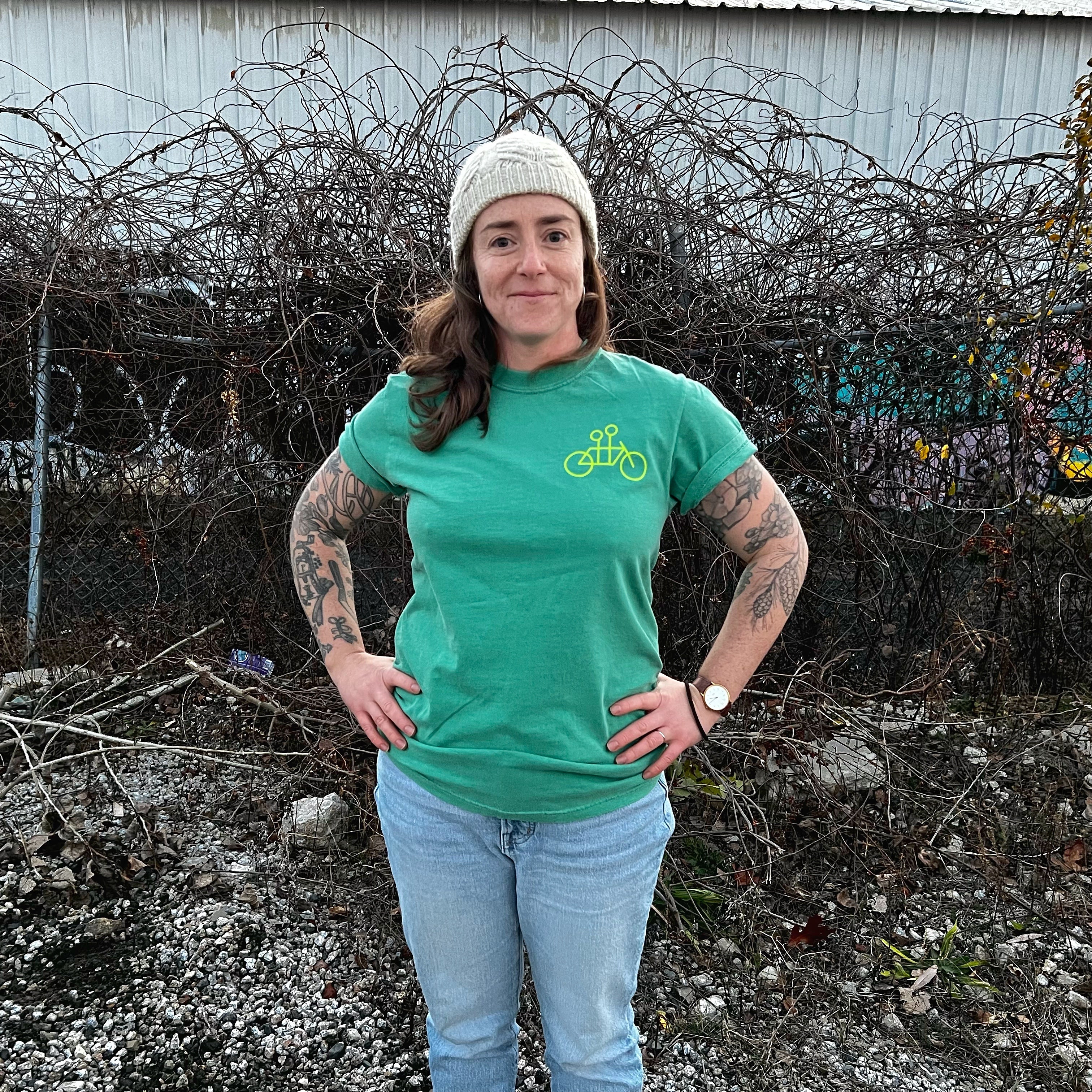 A woman in a Tandem Coffee Roasters tee and jeans stands confidently with hands on hips in front of a barbed wire fence, sporting a white beanie and tattoos on her arms.