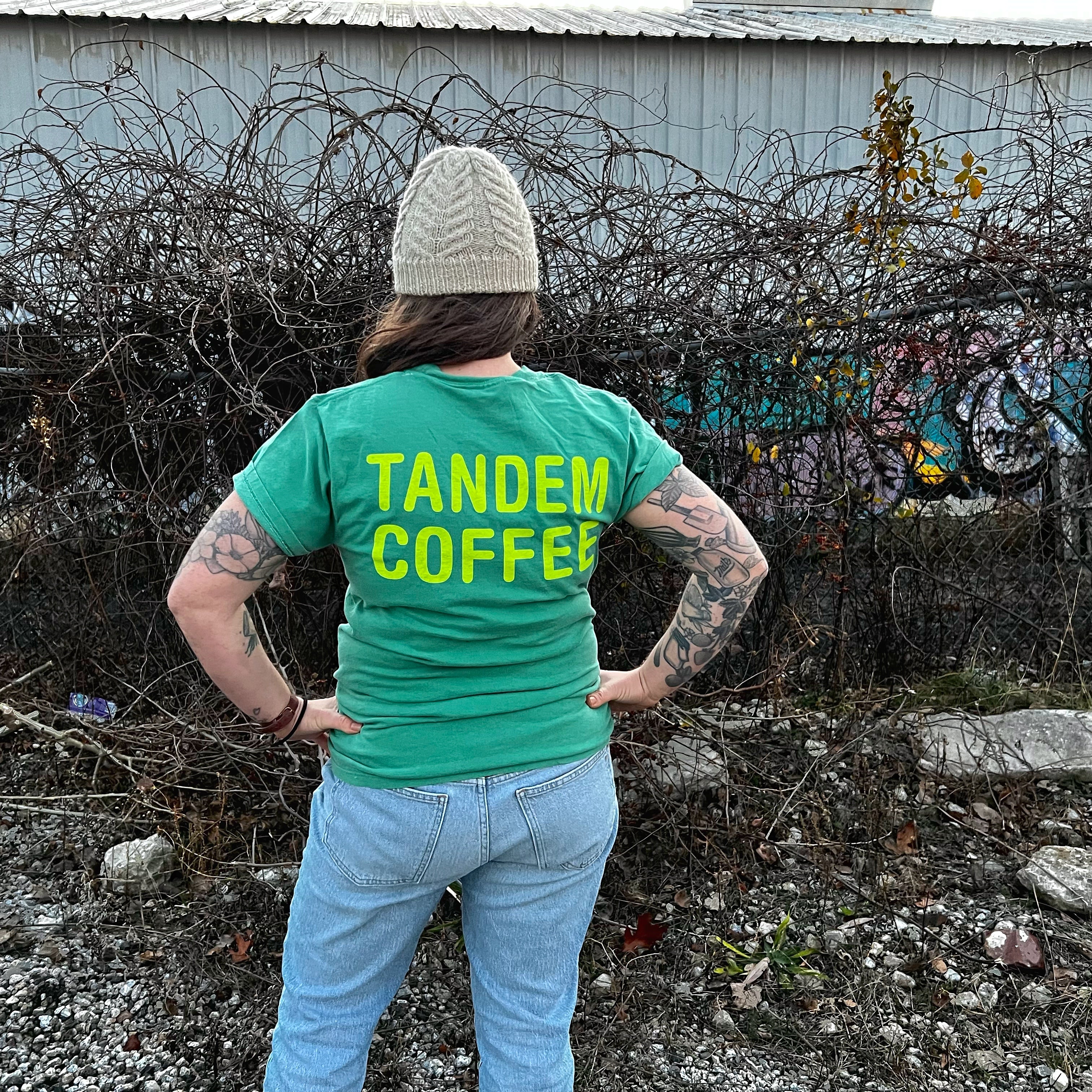 A person wearing a green "Tandem Coffee Roasters" logo tee and a beige beanie stands with hands on hips, facing away from the camera, with tattooed arms, in front of a graffiti