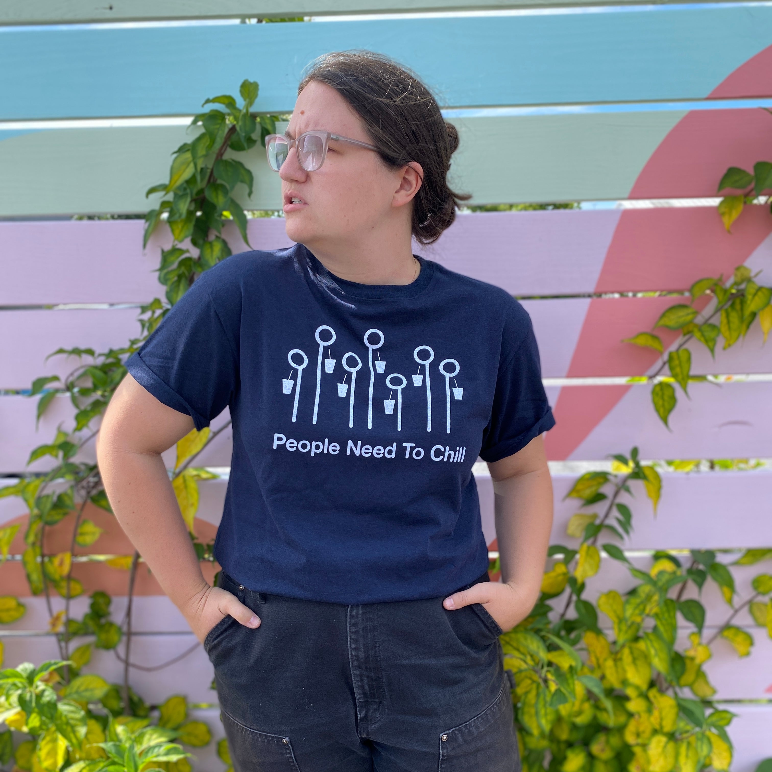 A person in a navy blue "People Need To Chill" Tee by Tandem Coffee Roasters, with pictograms, standing with hands on hips, looking to the side, against a colorful striped background.