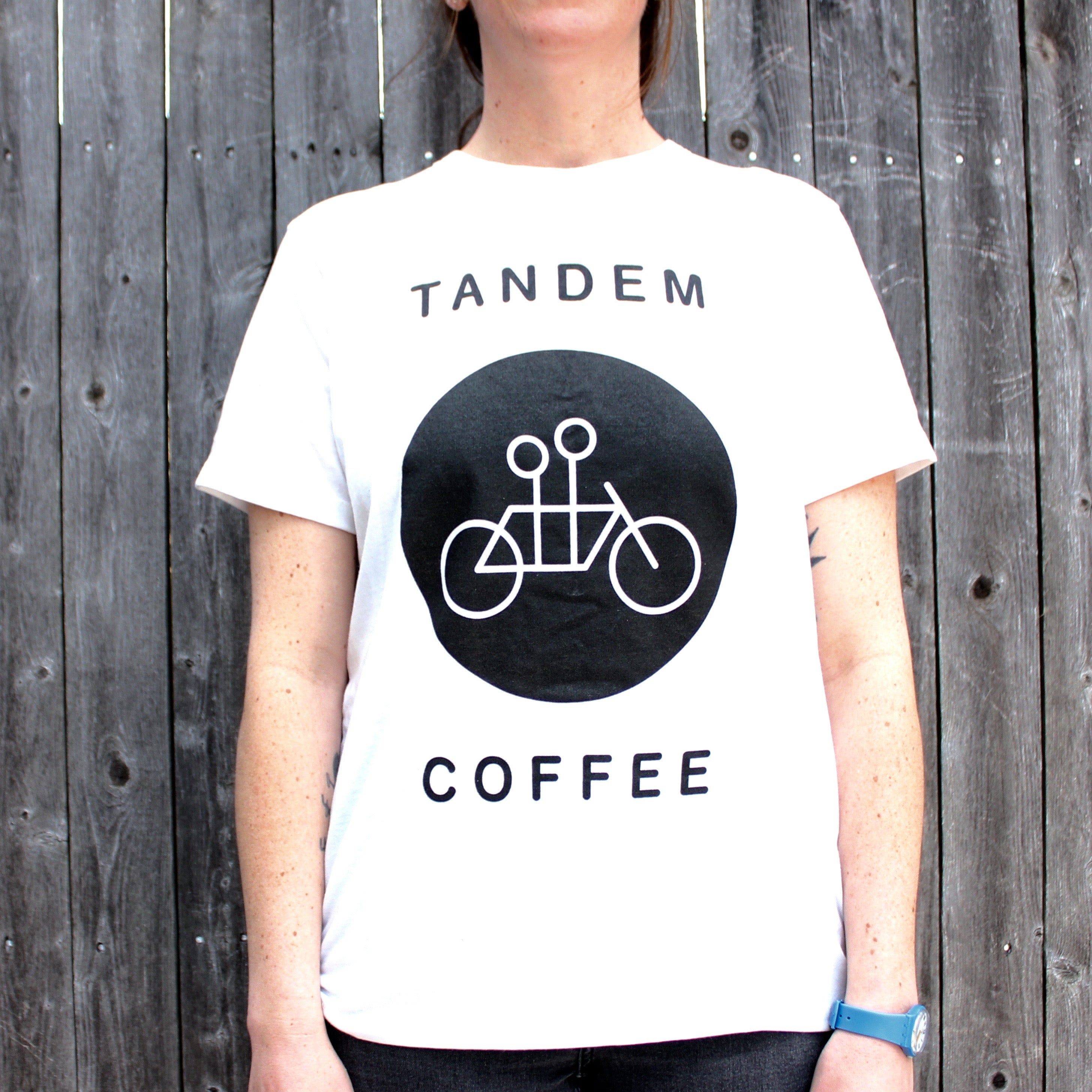 A person wearing a white Tandem Coffee Roasters Circle Logo Tee with a black circle design that includes a tandem bicycle illustration and the words "tandem coffee" standing against a wooden background.