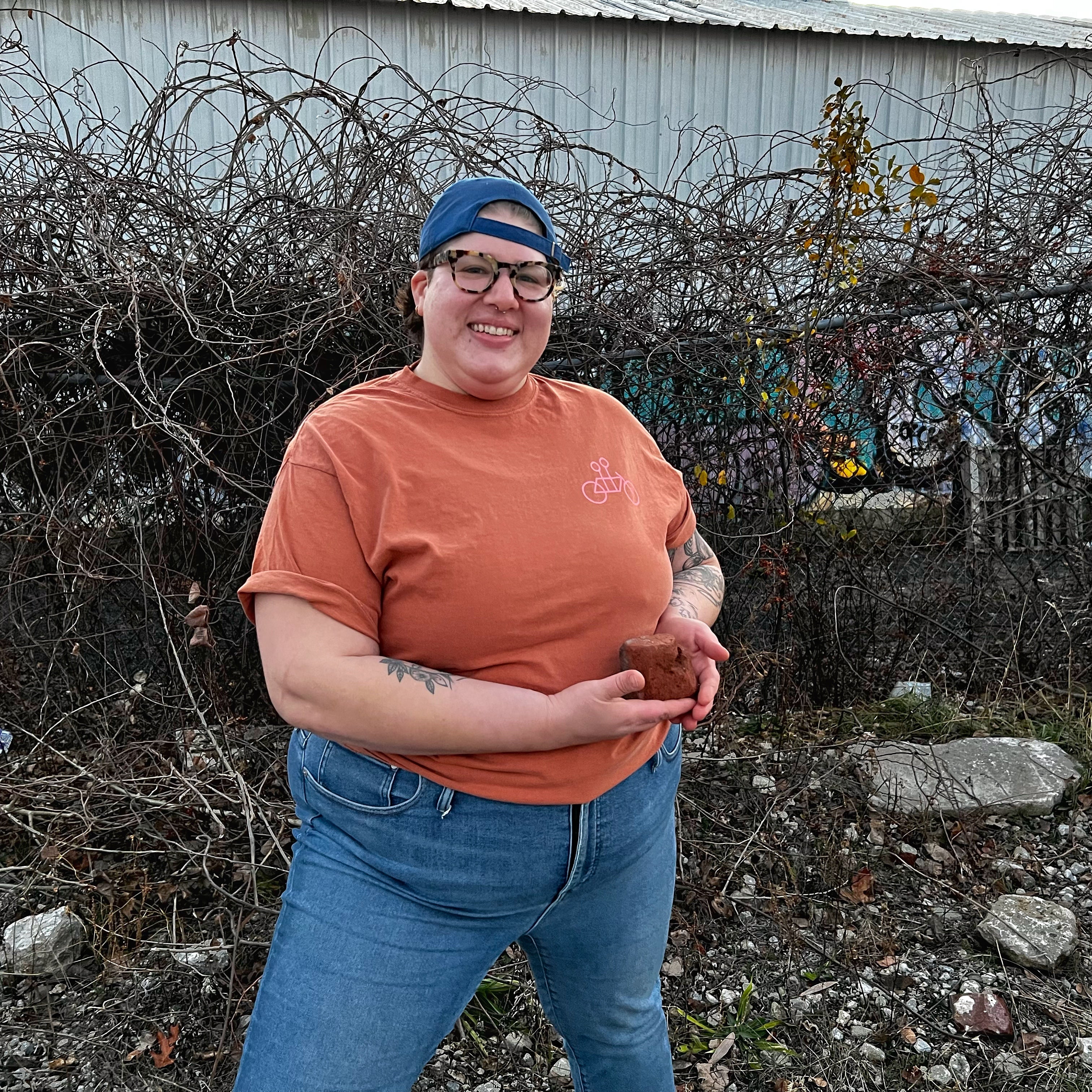 A person with a cap and glasses smiles while holding a Bike tee in terracotta, standing in front of a tangled, leafless bush and a white shed. They wear a Comfort Color tee in terracotta from Tandem Coffee Roasters.