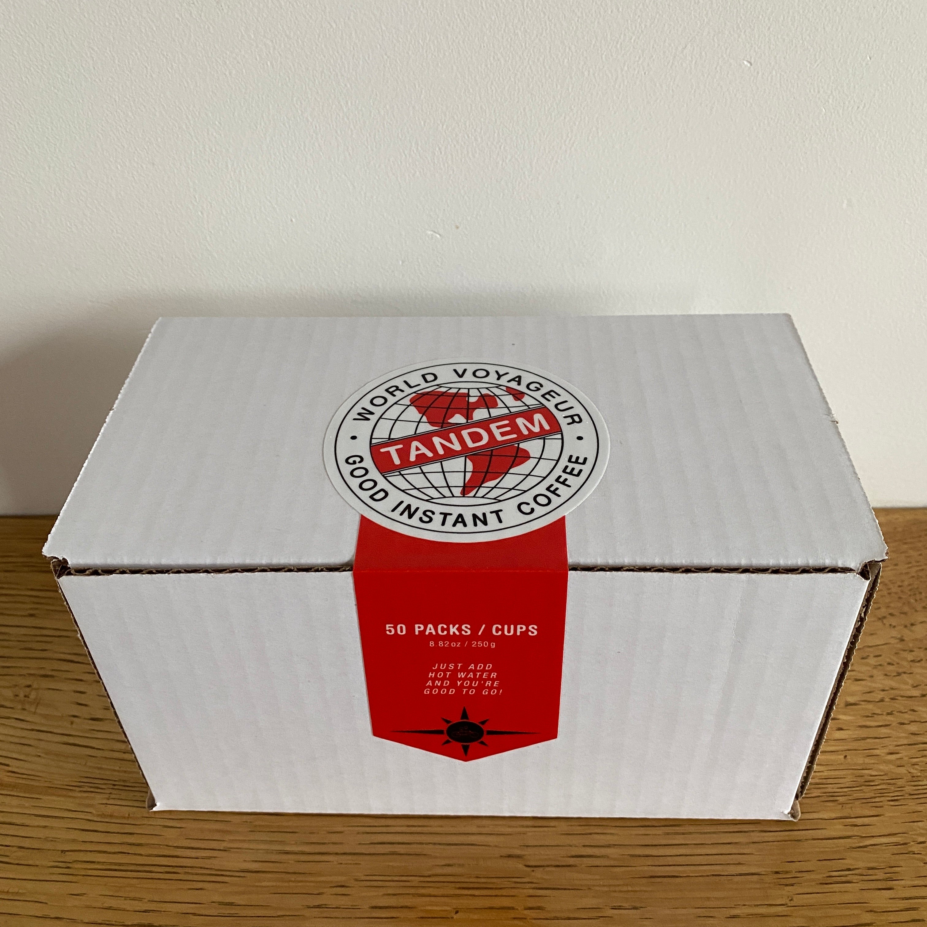 A white cardboard box with a red label sits on a wooden surface. The label reads "Tandem Coffee Roasters, Tandem World Voyageur - 50 Pack," and indicates the contents as "Superior Instant Coffee." It also features an image of a globe and compass accents, perfect for the jet-setting coffee lover.