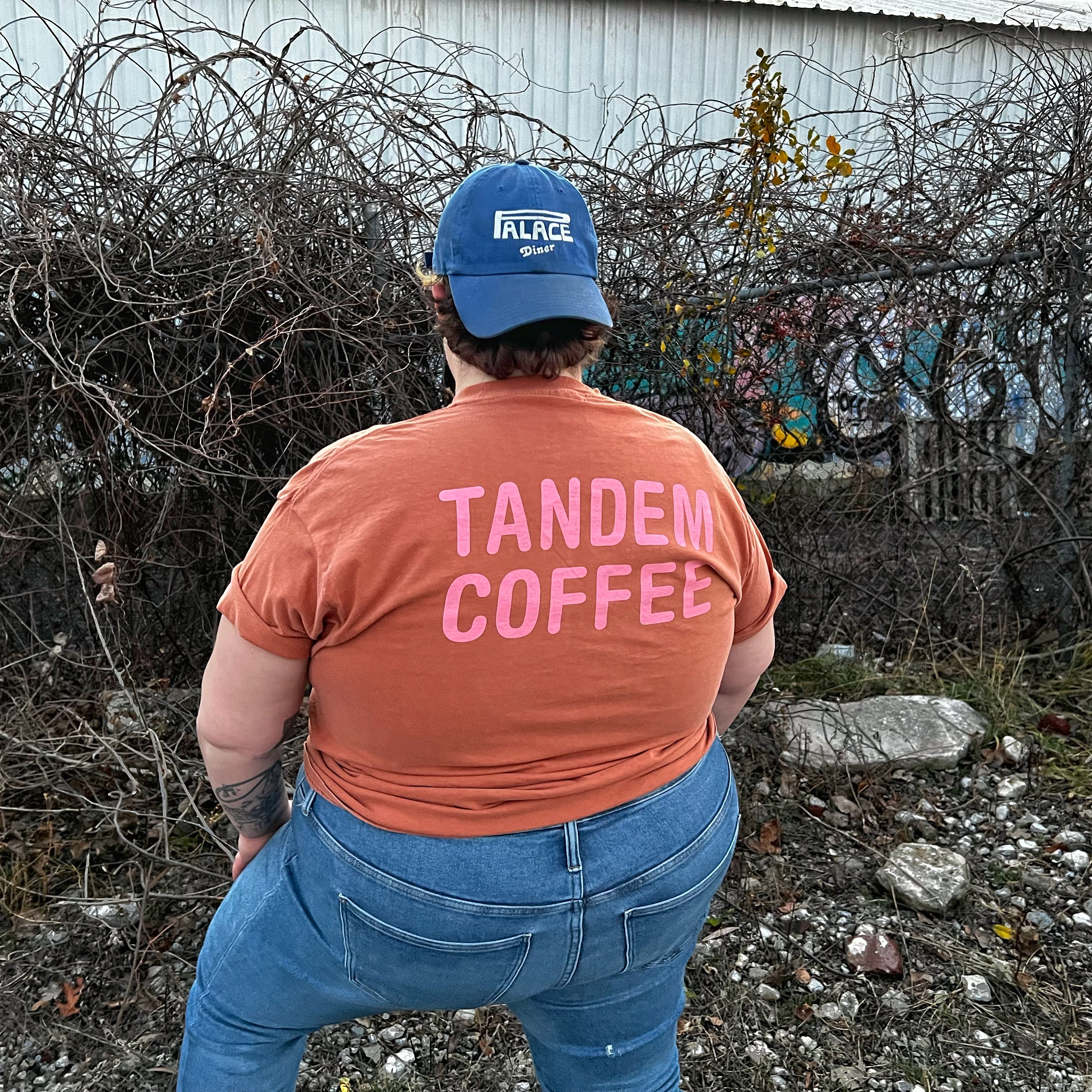 A person wearing a blue cap and a peach Comfort Color tee with "Tandem Coffee Roasters" written on the back, stands with their hands on hips facing a rustic wire fence and a graffiti-covered building.