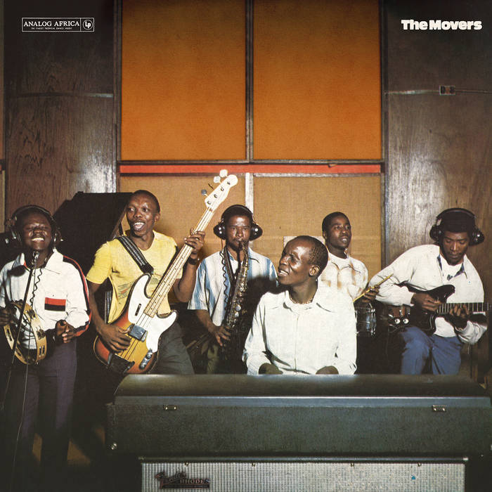 Six band members from The Movers - 1970 - 1976 are performing in a recording studio with wood-paneled walls, reminiscent of the Soweto Inn. The group includes a keyboardist in the foreground and musicians playing electric guitar, bass guitar, saxophone, drums, and another instrument. They appear to be enjoying themselves.